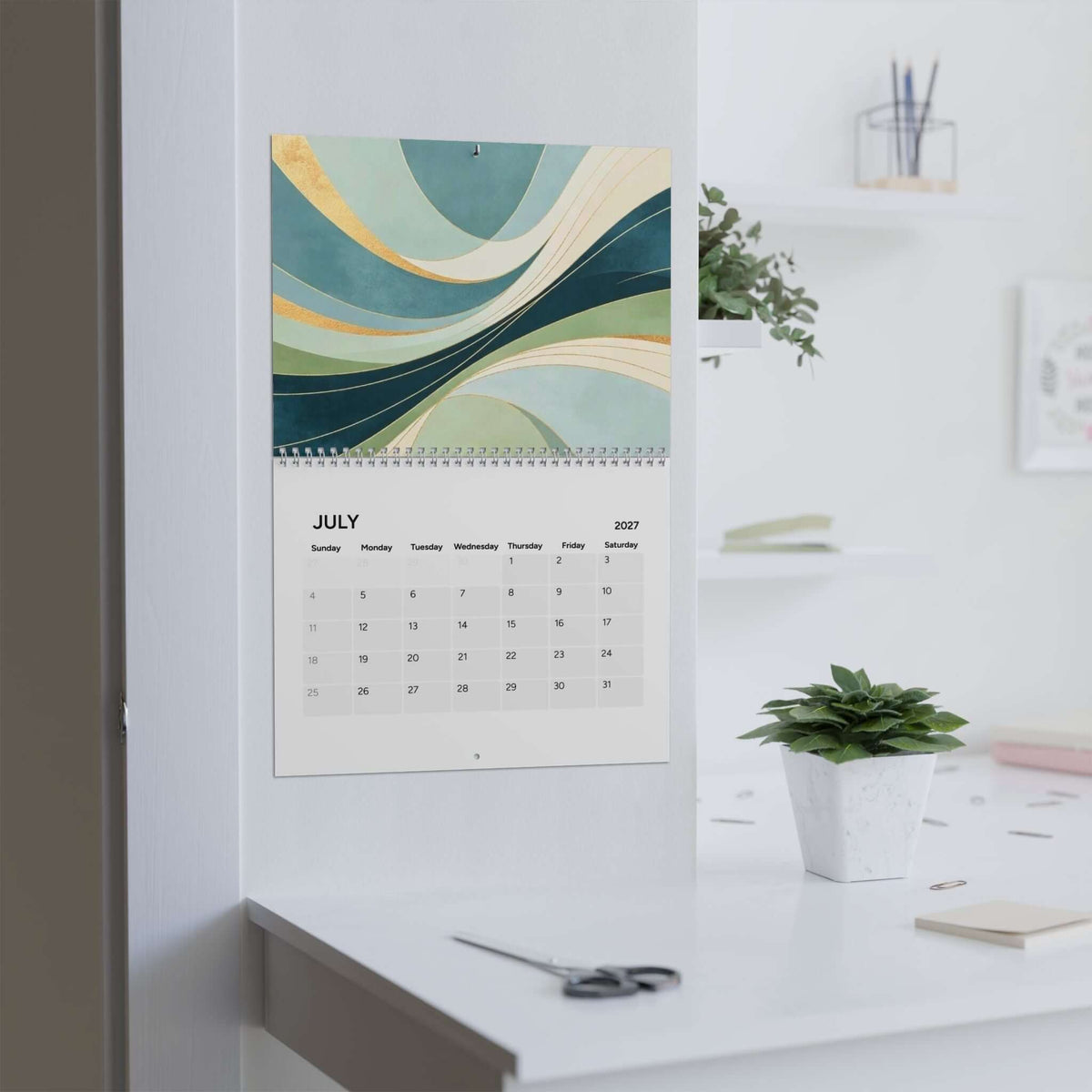 A wall-mounted 2027 July calendar with an abstract wave design in teal, green, cream, and gold, shown hanging on a white wall above a desk in a bright office setting.