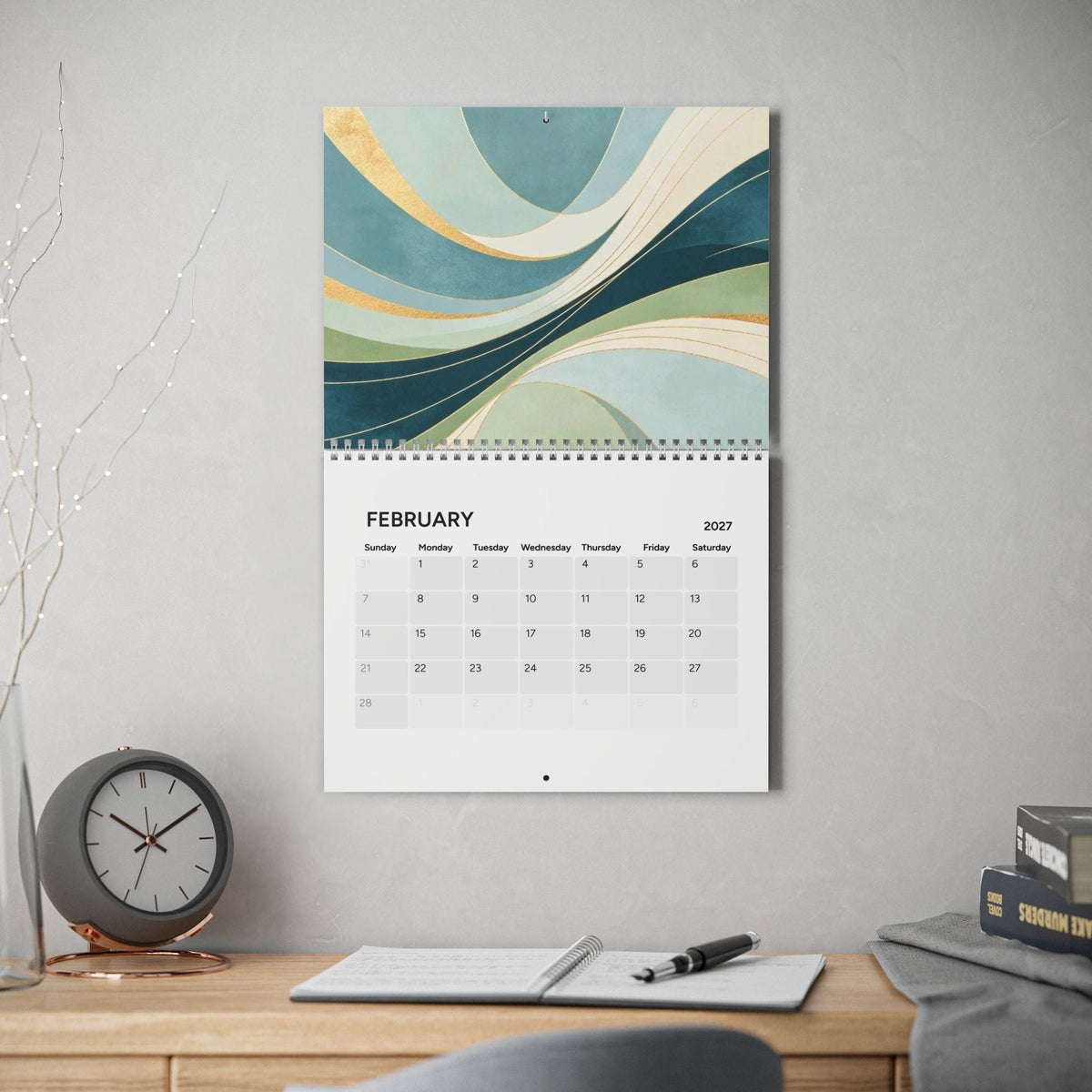 A hanging wall calendar page for February 2027 is displayed on a light gray wall above a wooden desk. The calendar features an abstract wave design in teal, blue, cream, and gold tones, with the rest of the page showing the monthly calendar grid.